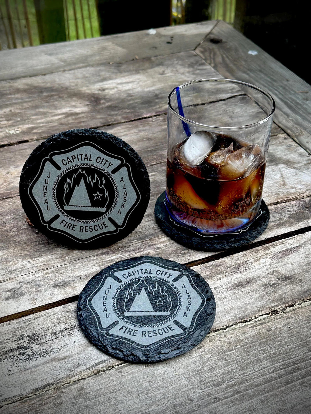 Two slate coasters with 'Capital City Fire Rescue' design on a wooden surface with a glass of iced coffee.